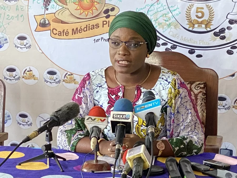 Femme en tenue colorée et foulard vert qui parle face à plusieurs micros lors d’une conférence de presse