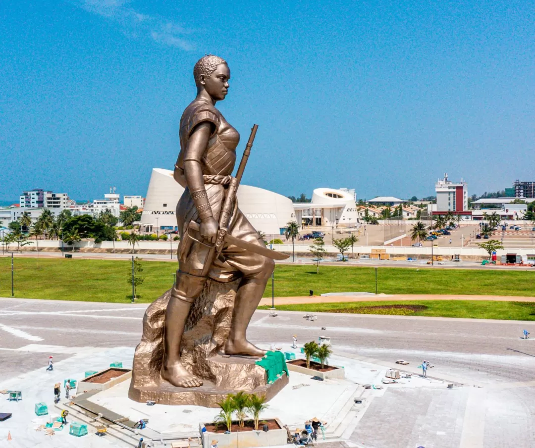 Statue monumentale d’une femme tenant un bâton sur une grande place avec bâtiments modernes en arrière-plan