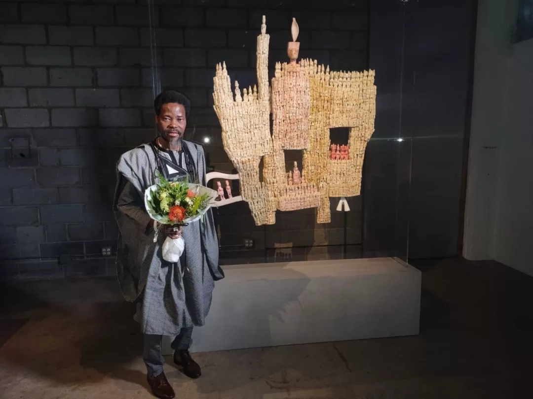 Un homme en tenue traditionnelle tient un bouquet près d'une sculpture en forme de main ornée de petites figurines en exposition