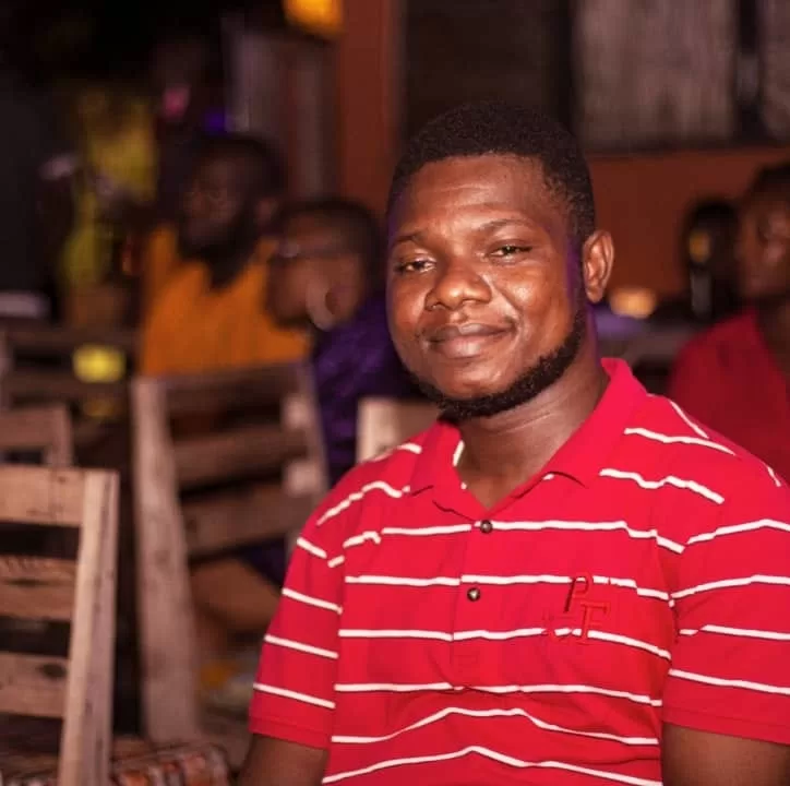 Homme souriant portant un polo rouge à rayures blanches assis dans un lieu intérieur avec des chaises en bois autour