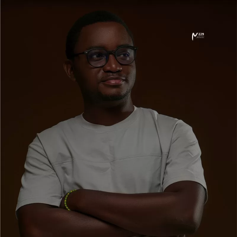 Homme portant des lunettes et un t-shirt clair, bras croisés, regard dévié, fond sombre