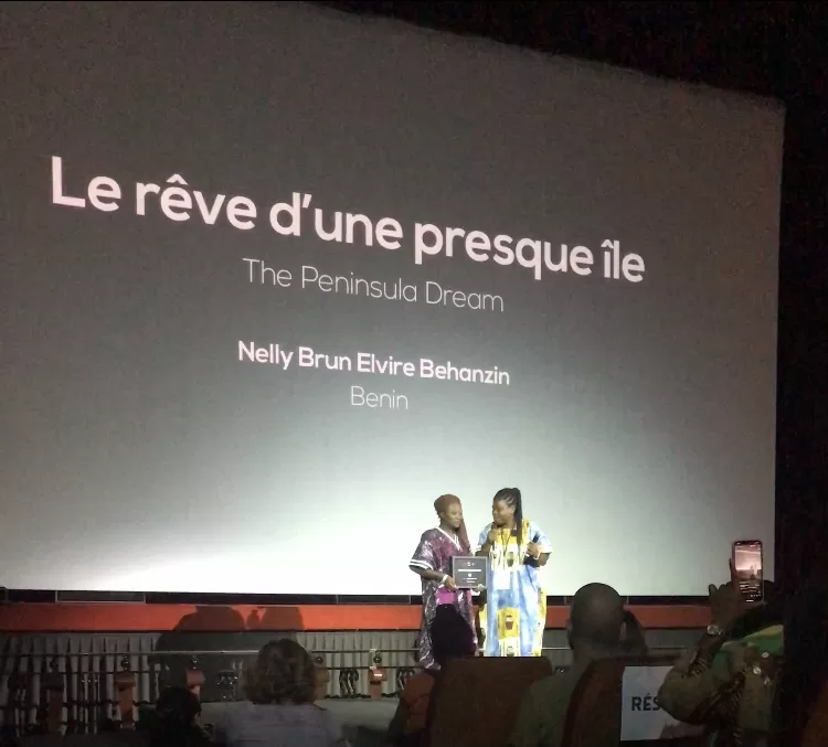 Deux personnes sur scène devant un grand écran portant le titre d’un film et un nom avec un public assis au premier plan