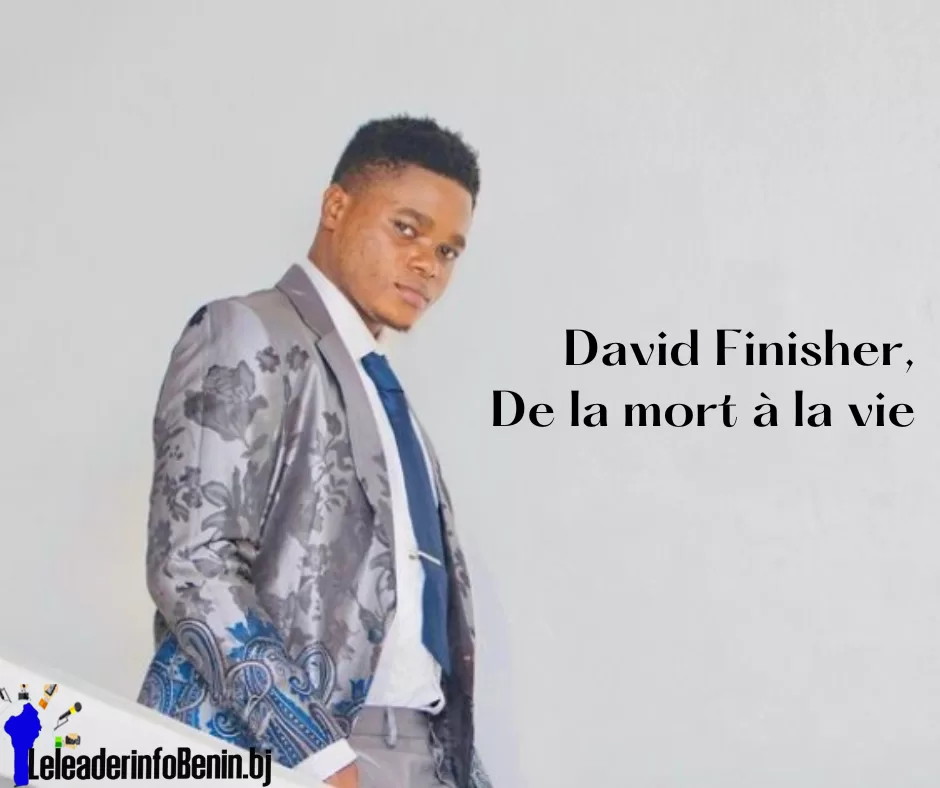 Jeune homme en costume gris à motifs floraux avec cravate bleue posé devant un mur blanc avec texte en noir à côté