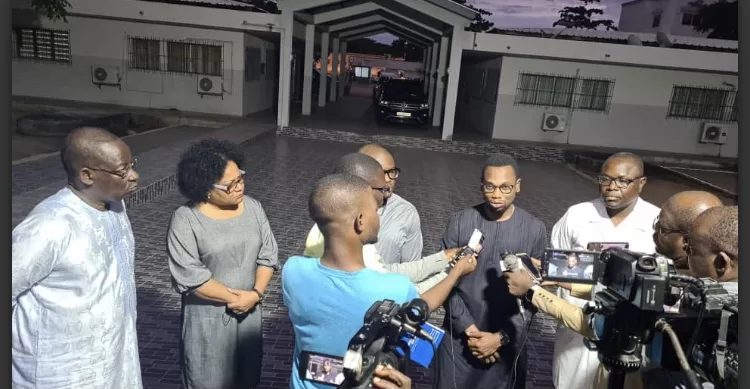 Un groupe de personnes discute devant des microphones et des caméras dans une cour extérieure au crépuscule