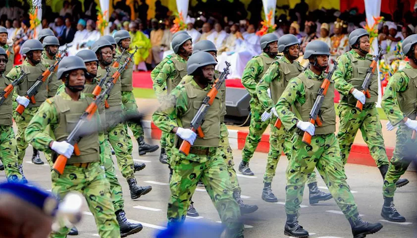 Des soldats en tenue camouflage verte et casques défilent dans une rue devant des spectateurs assis sous un chapiteau coloré