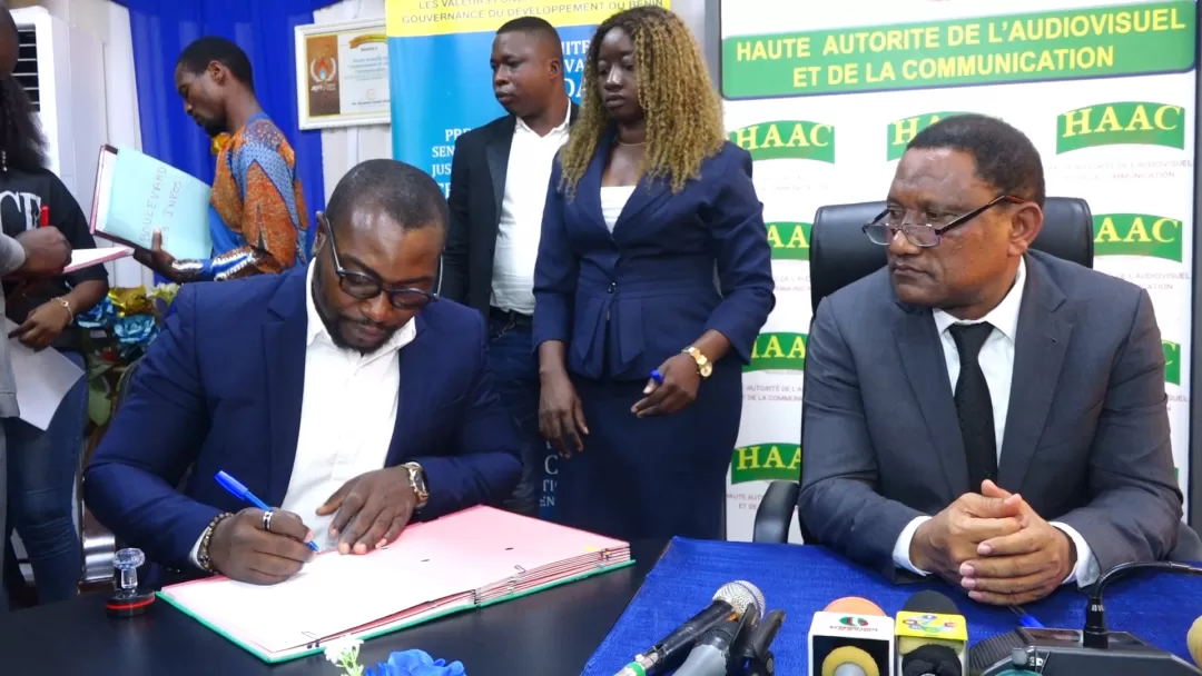 Un homme signe un document à une table tandis qu'un autre homme en costume sombre et trois personnes posent en arrière-plan devant des panneaux HAAC