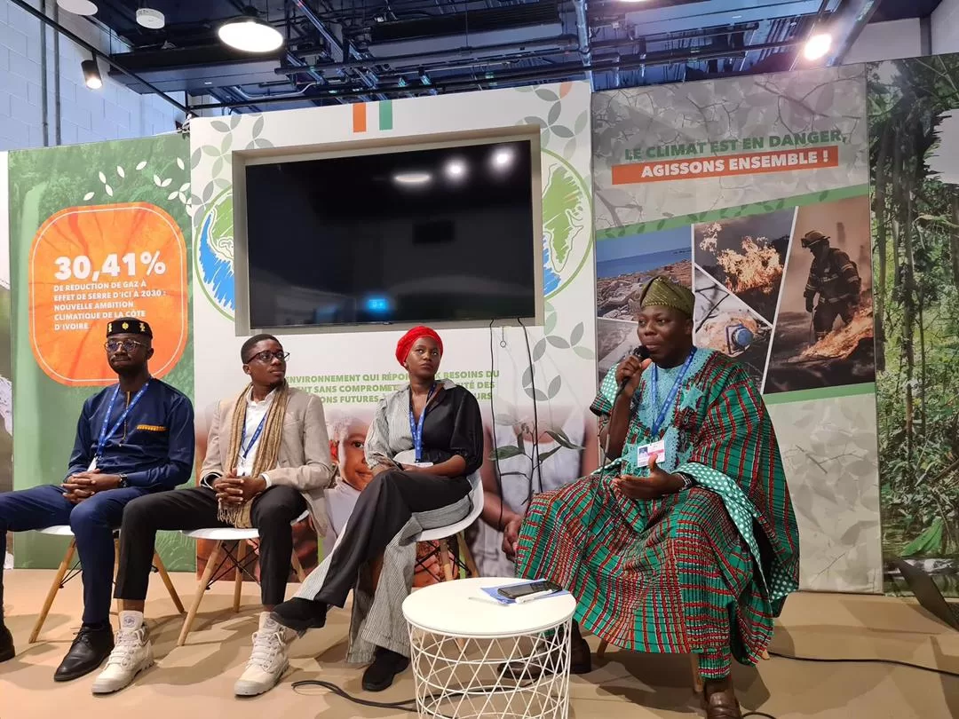 Quatre personnes assises en discussion avec une femme en tenue traditionnelle tenant un micro dans un espace d’exposition sur le climat