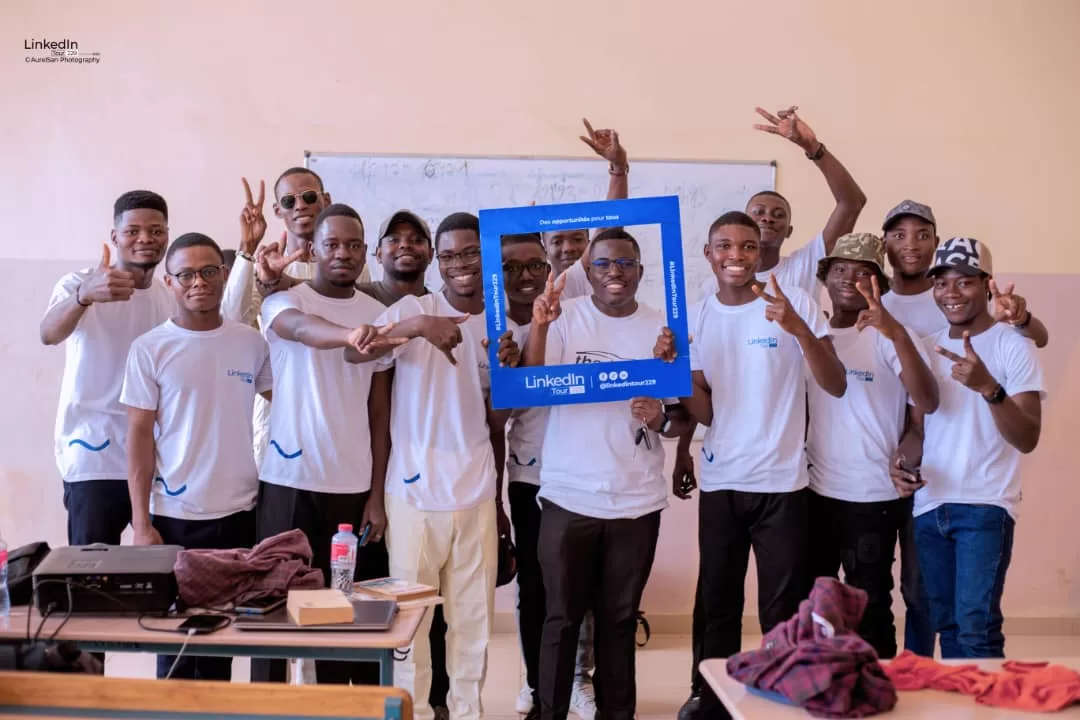 Un groupe de jeunes hommes souriants portent des t-shirts blancs et font des gestes de victoire en posant avec un cadre LinkedIn bleu dans une salle de classe