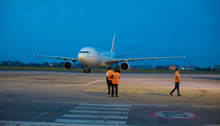 Avion stationné sur la piste d’aéroport avec trois personnels en gilets orange au premier plan au crépuscule