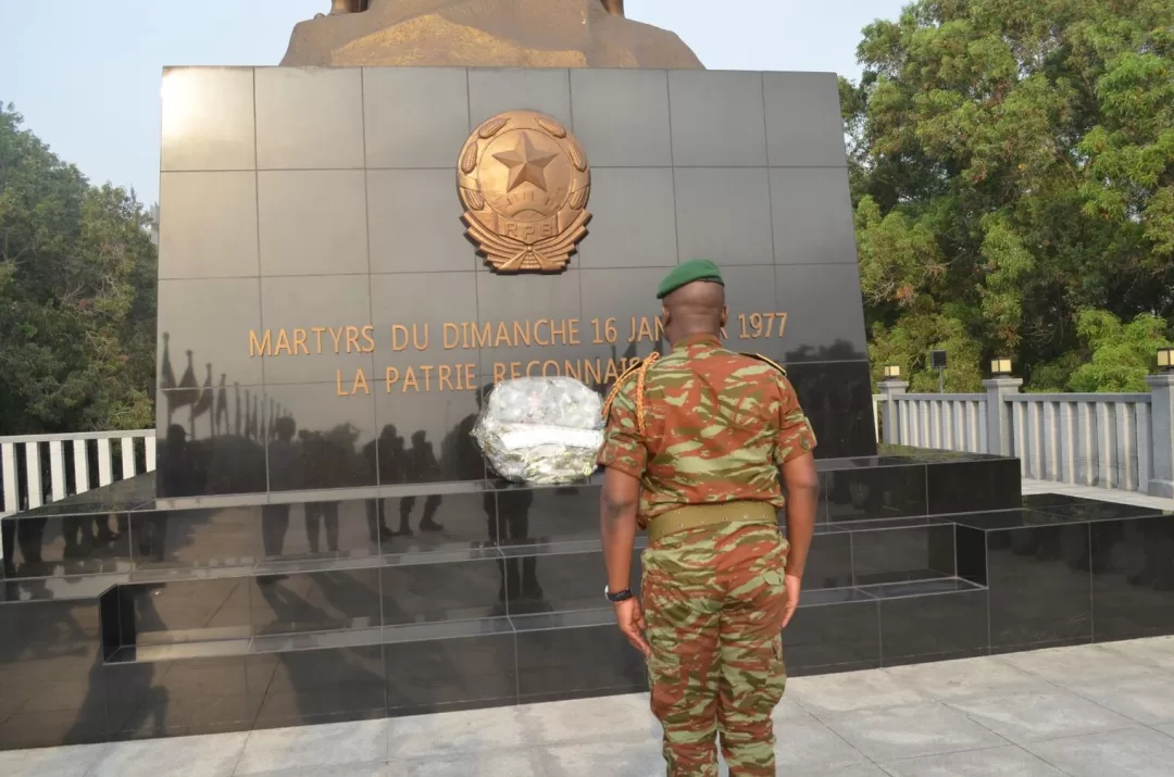 Un militaire en tenue camouflage et béret vert face à un monument commémoratif orné d’une gerbe de fleurs blanches