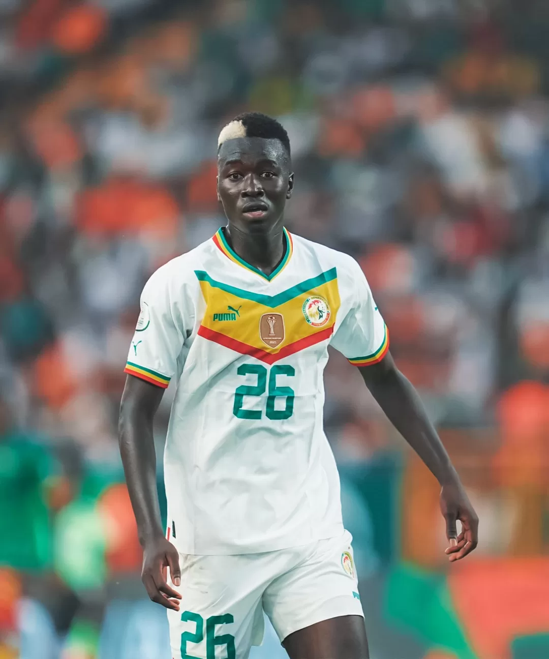 Un joueur de football en maillot blanc avec le numéro 26 sur un terrain avec des spectateurs flous en arrière-plan