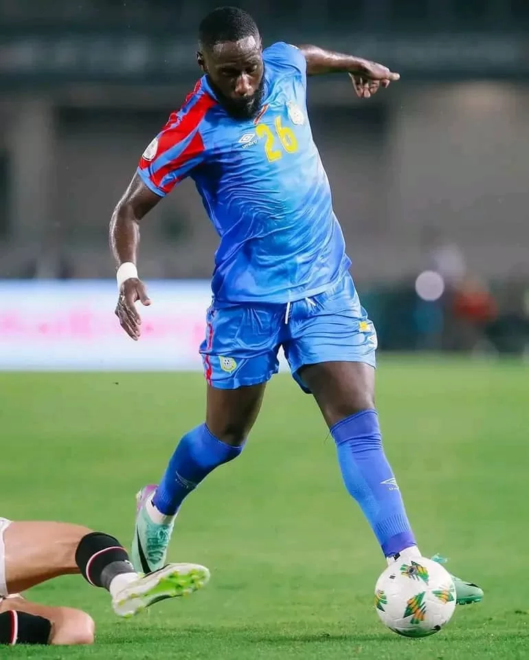 Joueur en maillot bleu avec numéro 26 contrôle le ballon face à un adversaire au sol lors d’un match de football