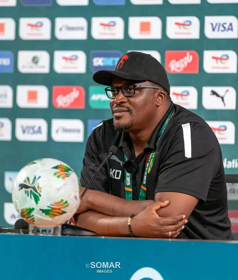 Un homme en polo noir et casquette noire avec lunettes parle lors d’une conférence avec un ballon de football sur la table devant lui