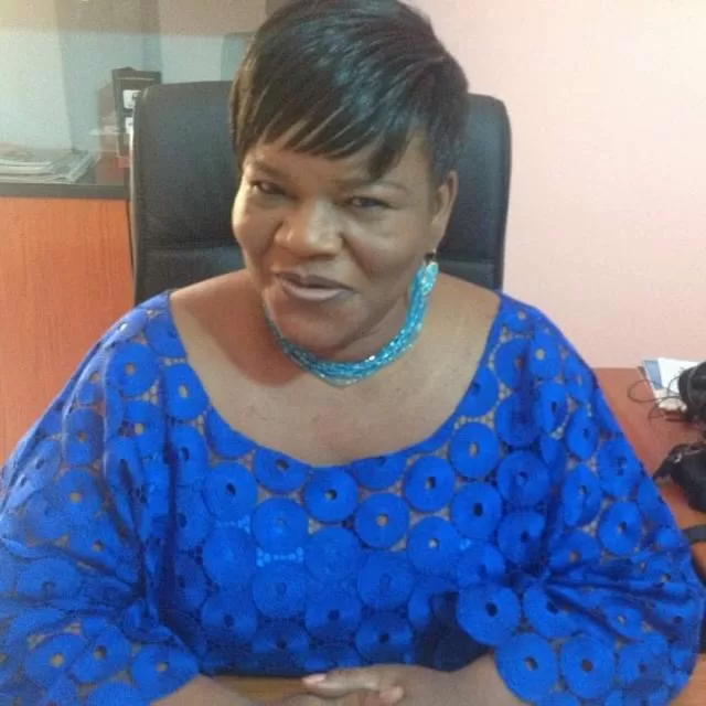 Femme souriante en tenue traditionnelle bleue assise dans un bureau devant un meuble et un ordinateur.