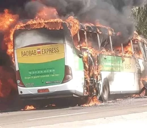 Un bus est entièrement en flammes, avec de la fumée noire s'échappant du toit et des côtés sur une route.