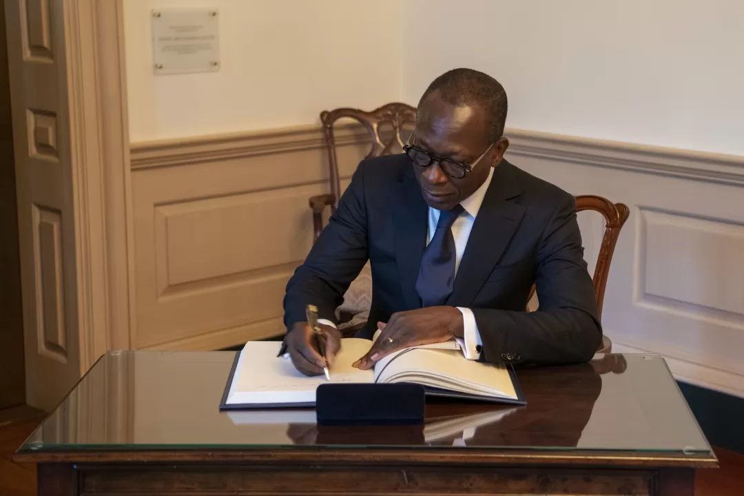 Un homme en costume sombre signe un document posé sur une table en bois dans une pièce à la décoration classique
