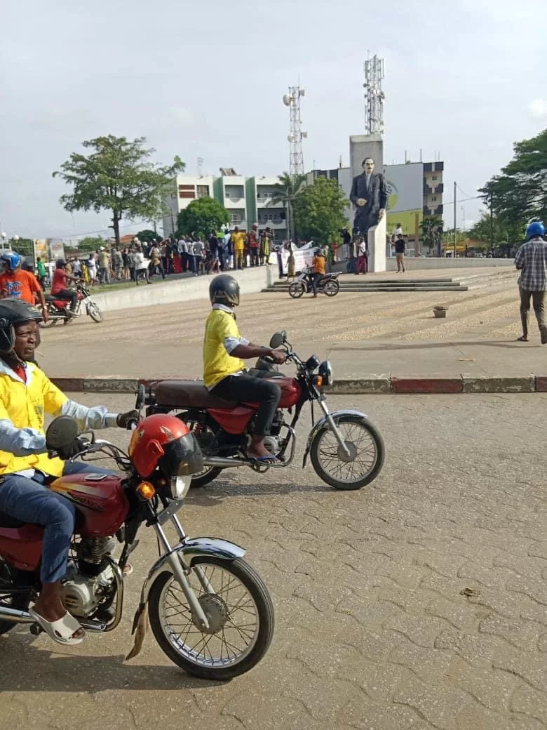 Deux hommes en moto passent devant une place avec une foule rassemblée près d'une statue imposante