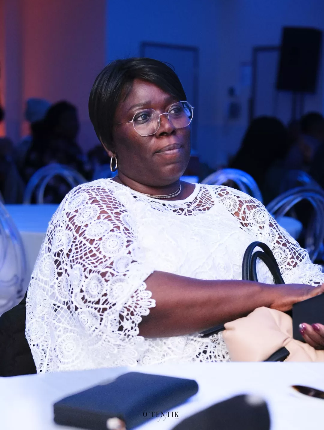Femme portant des lunettes et un haut blanc en dentelle assise à une table lors d’un événement en intérieur
