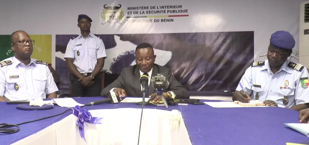 Quatre hommes en uniformes et costume assis à une table avec microphones et documents devant un panneau du ministère de l’Intérieur et de la Sécurité publique