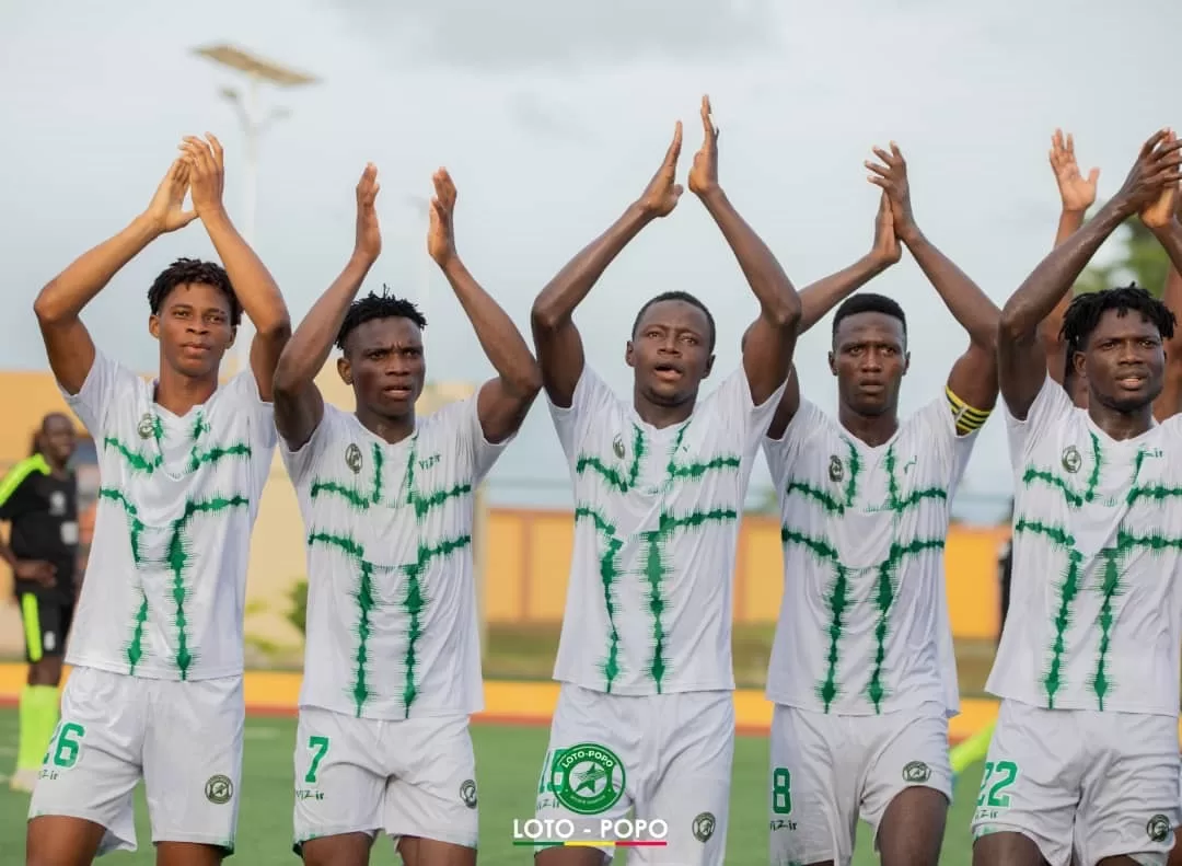 Six joueurs de football en maillot blanc à motifs verts applaudissent côte à côte sur un terrain de jeu.