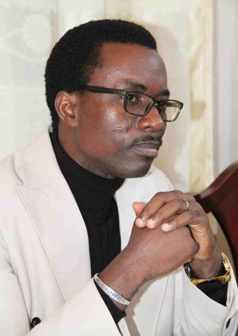 Homme portant des lunettes et un pardessus clair, les mains jointes, regard pensif vers la droite