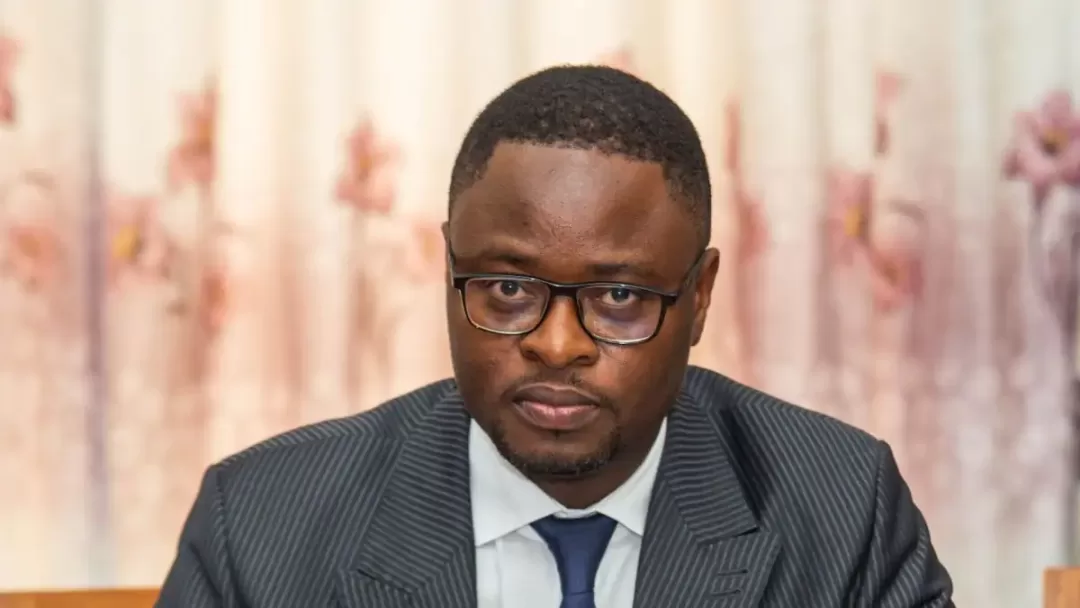 Homme portant des lunettes et un costume rayé avec cravate assis devant un rideau fleuri rose pâle