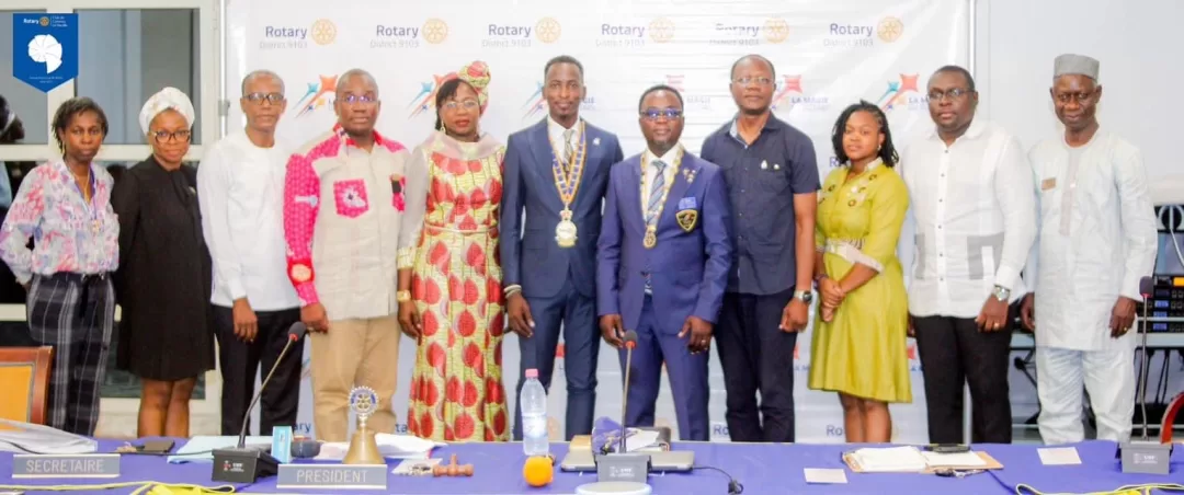 Dix personnes debout en rang devant une table avec des microphones et des documents dans une salle de réunion décorée de logos Rotary