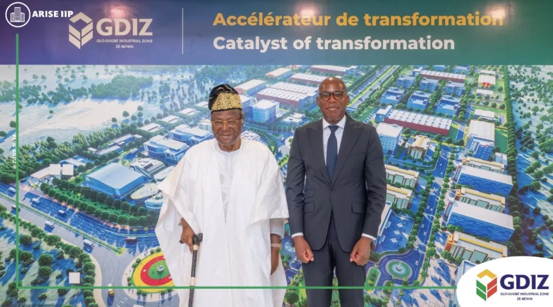 Deux hommes debout, l’un en tenue traditionnelle blanche et l’autre en costume sombre, devant une grande image aérienne colorée d’une zone industrielle