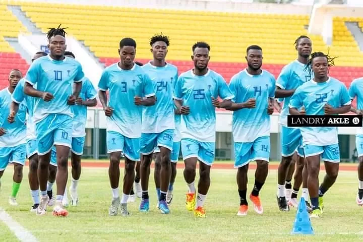 Un groupe de joueurs de football en tenue bleu clair s'entraîne en courant sur un terrain devant des gradins colorés