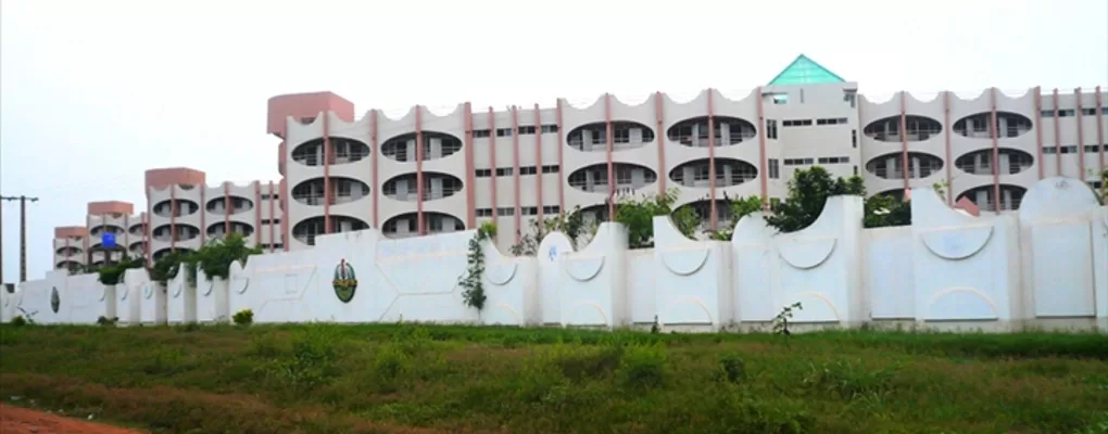 Grand bâtiment moderne blanc avec plusieurs étages derrière un mur blanc partiellement décoré et un espace herbeux au premier plan