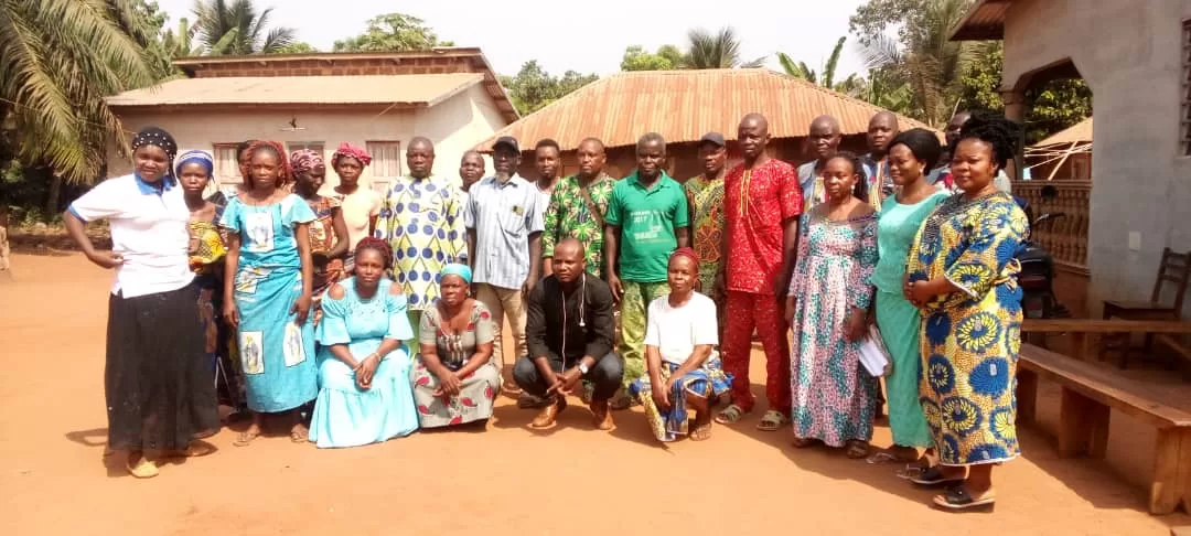 Groupe d’hommes et de femmes posant devant des maisons traditionnelles en milieu rural sous un ciel clair