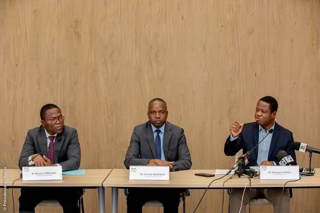Trois hommes en costume assis derrière une table lors d'une réunion formelle avec microphones et documents posés devant eux