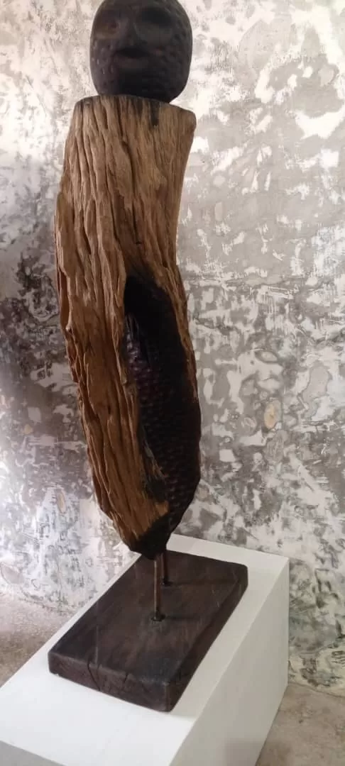 Sculpture en bois partiellement brûlé montée sur un socle blanc devant un mur texturé clair