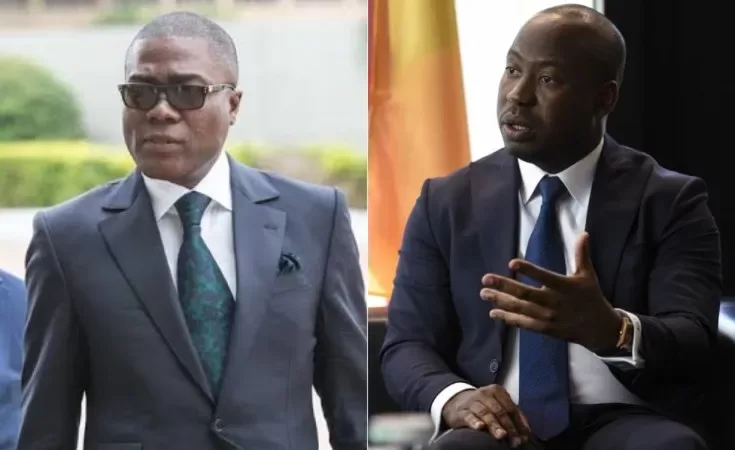 Deux hommes en costume formel, l’un avec lunettes noires, l’autre en pleine conversation avec geste de la main
