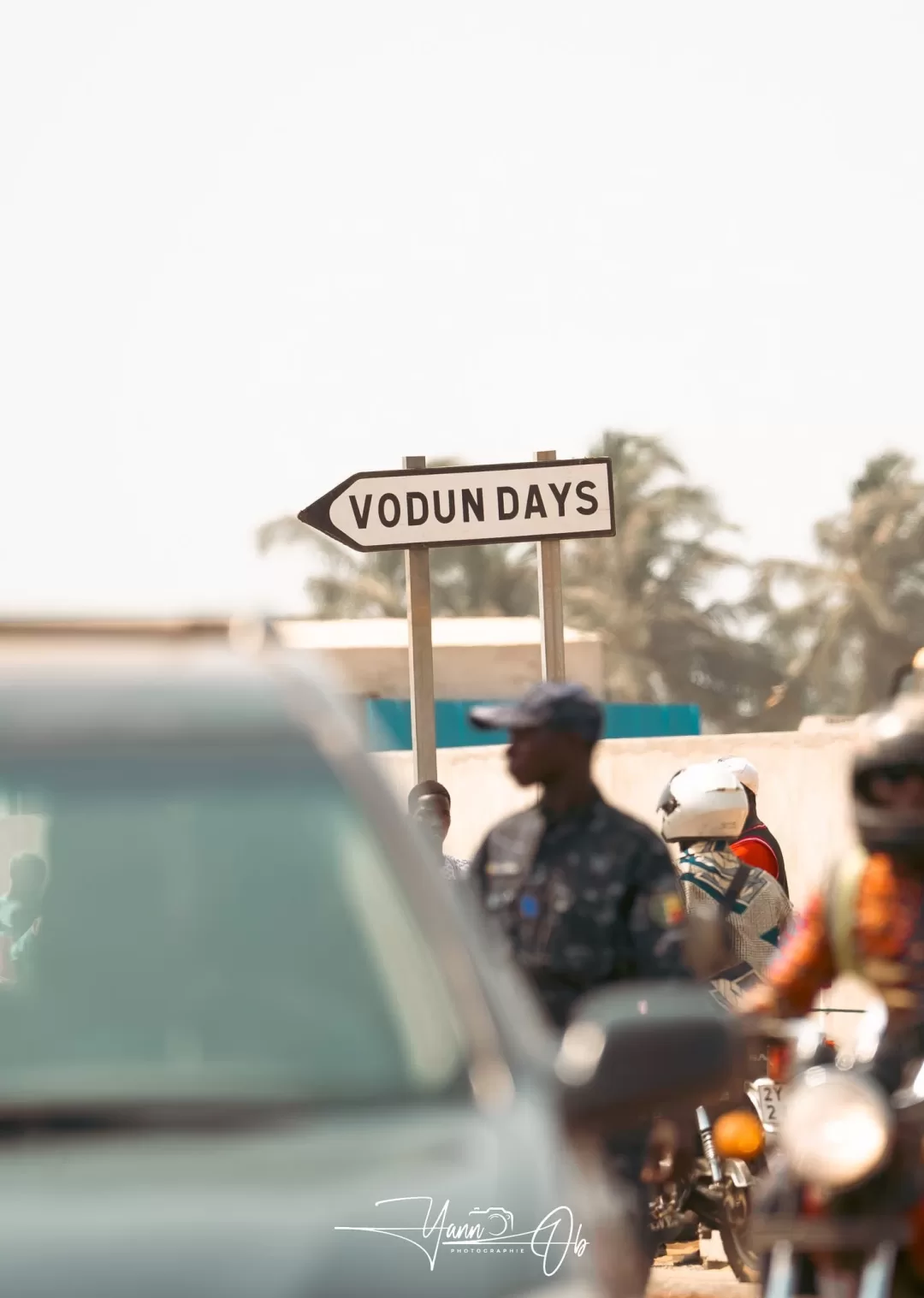Panneau directionnel « VODUN DAYS » tenu au-dessus d'un trottoir avec des passants et des véhicules flous au premier plan