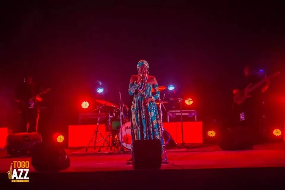 Chanteuse en tenue traditionnelle colorée sur scène éclairée en rouge avec musiciens en arrière-plan et logo Togo Jazz visible
