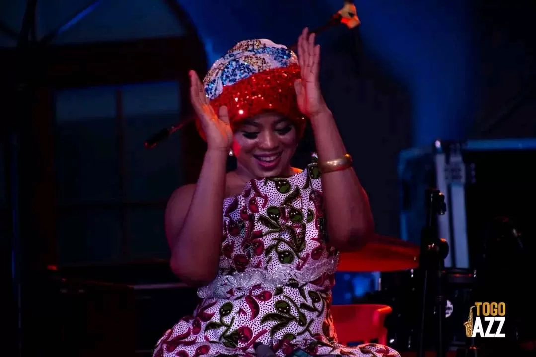 Femme souriante en tenue traditionnelle colorée et coiffe chantant et dansant sur scène éclairée en bleu et rouge