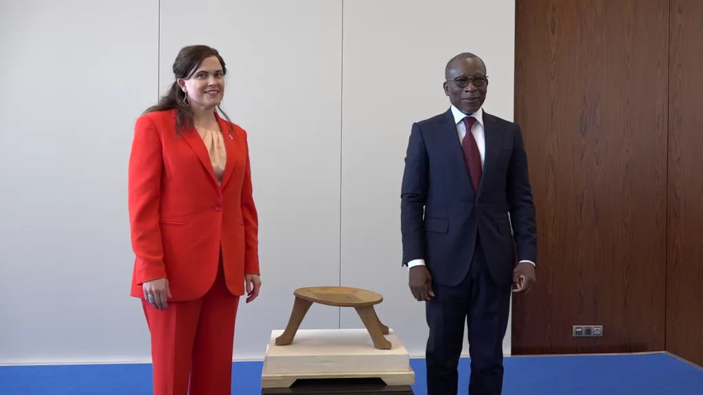 Un homme en costume sombre et une femme en tailleur rouge posent debout près d’un petit tabouret en bois sur un socle beige