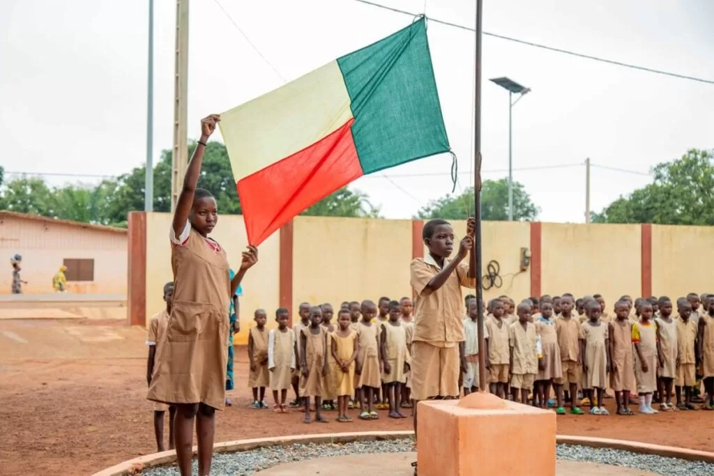 Des élèves en uniforme participent à une cérémonie de lever de drapeau devant un groupe d’enfants alignés dans une cour d’école