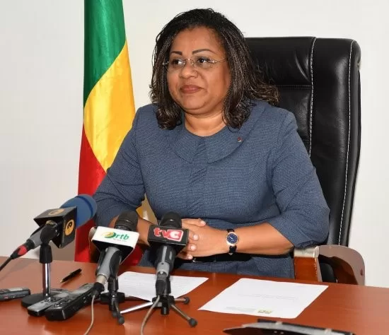 Une femme assise derrière un bureau avec plusieurs microphones et un drapeau aux couleurs vert, jaune et rouge en arrière-plan