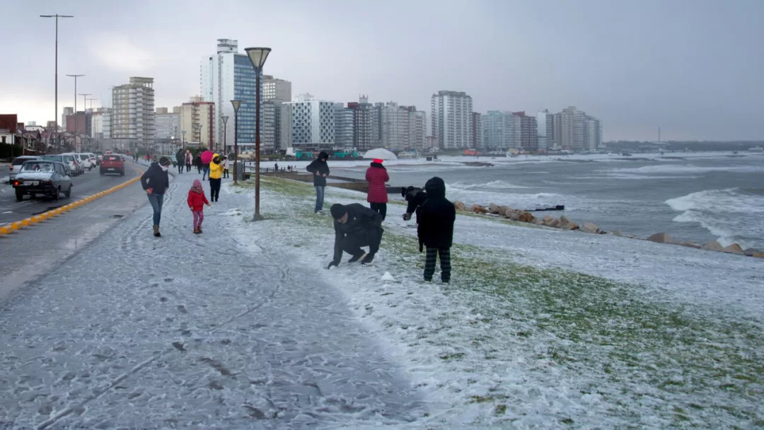 Personnes marchant et jouant sur un sol partiellement couvert de neige près d'une plage et de bâtiments en bord de mer