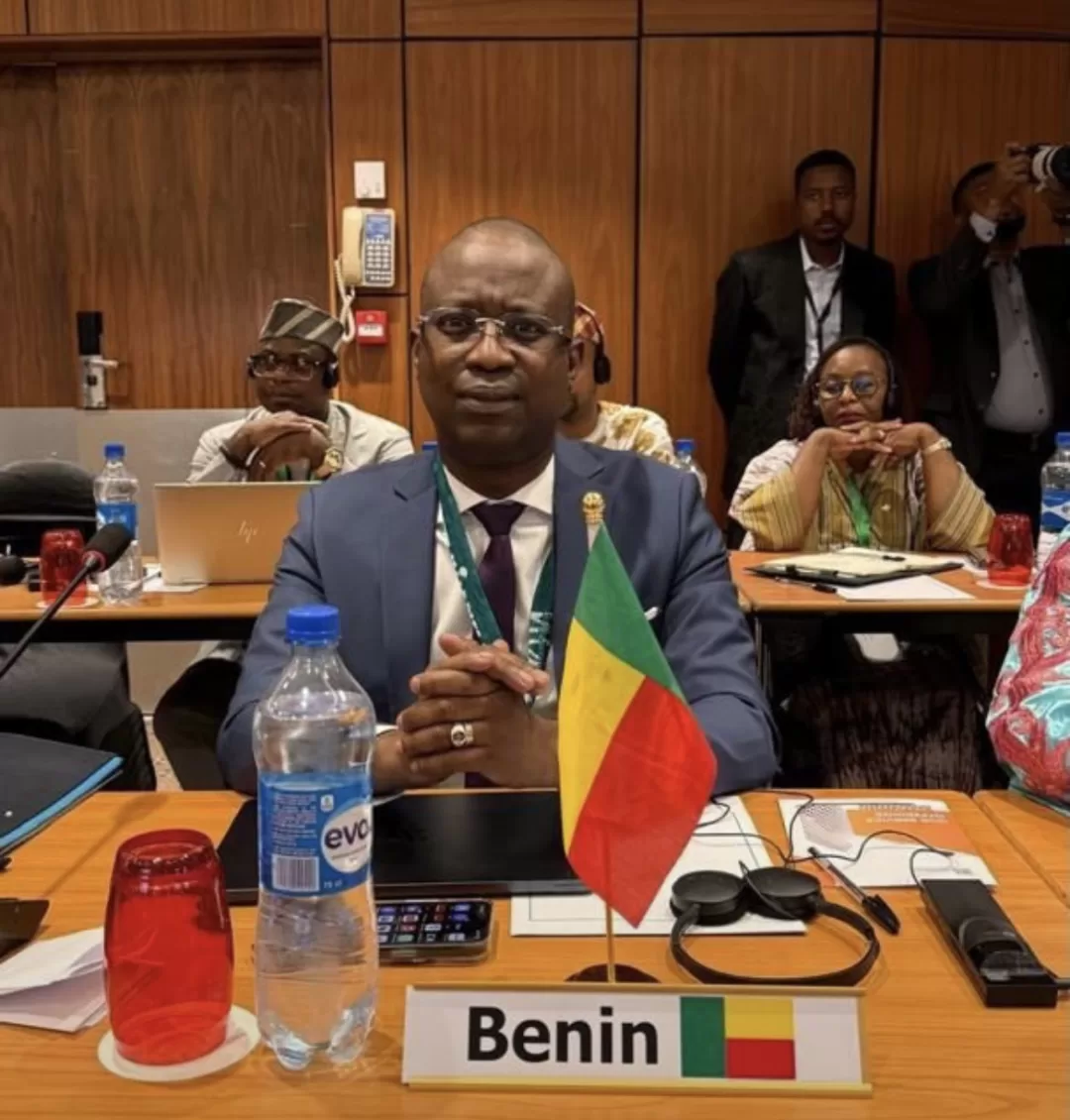 Homme en costume assis derrière une table avec un drapeau et une pancarte Benin entouré de plusieurs personnes dans une salle de réunion