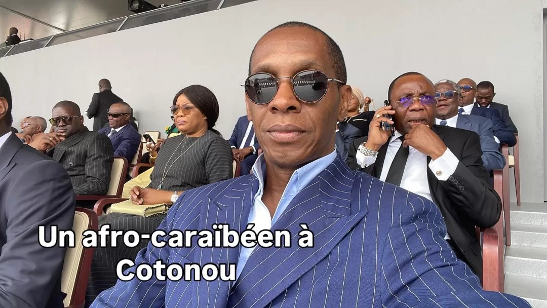 Un homme en veste rayée bleue et lunettes noires assis dans une tribune avec plusieurs autres personnes derrière lui