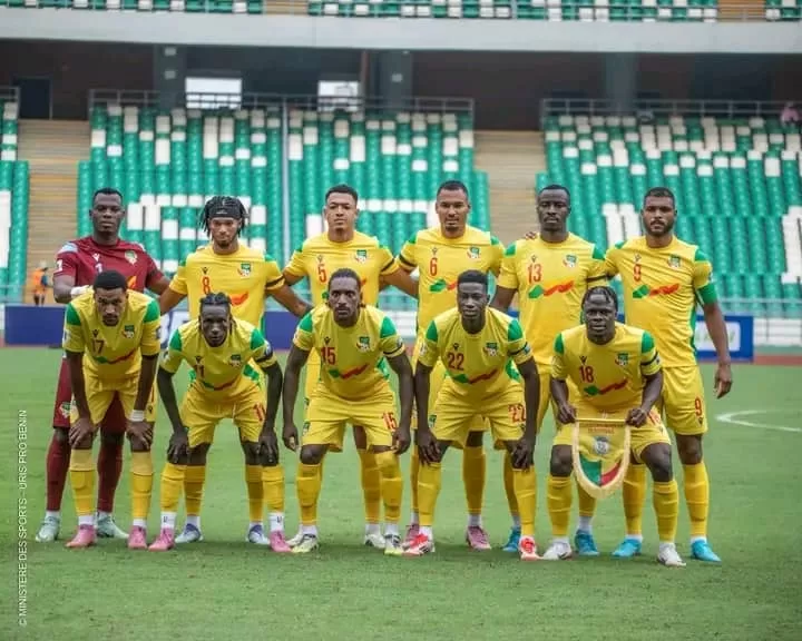 Onze joueurs de football en tenue jaune posent sur un terrain dans un stade avec des sièges verts et blancs vides