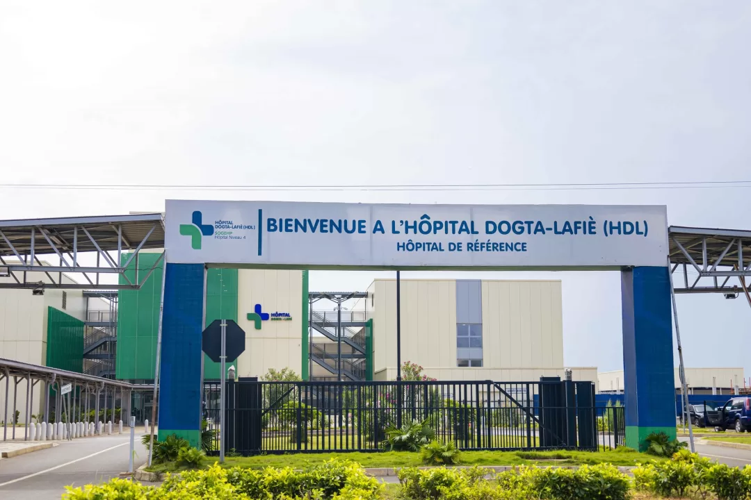 Entrée principale d’un hôpital moderne avec un portail, une barrière fermée et des plantes au premier plan