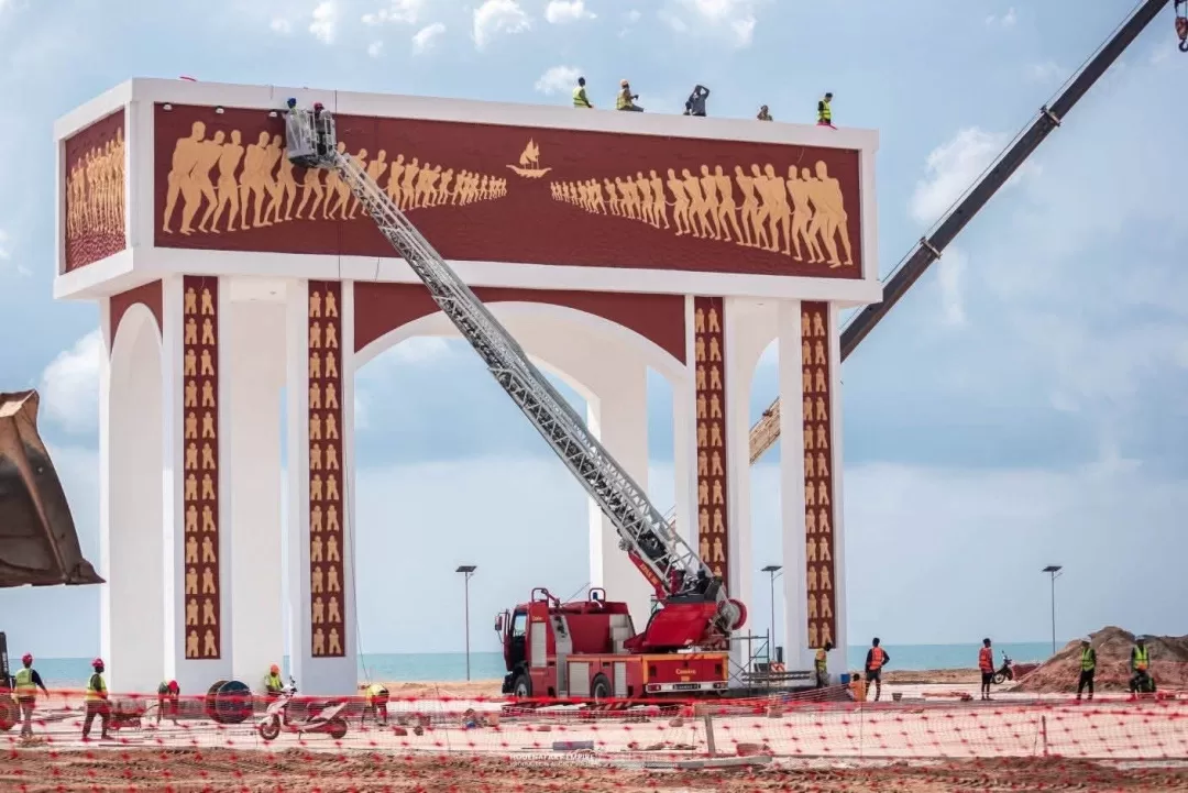 Grand portail décoré de silhouettes humaines en construction au bord de la mer avec des ouvriers et un camion-grue rouge