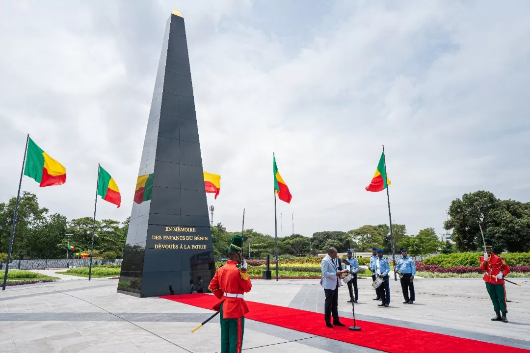 Un monument noir entouré de drapeaux tricolores rouges, jaunes, verts avec des militaires en uniforme et un homme au micro sur tapis rouge