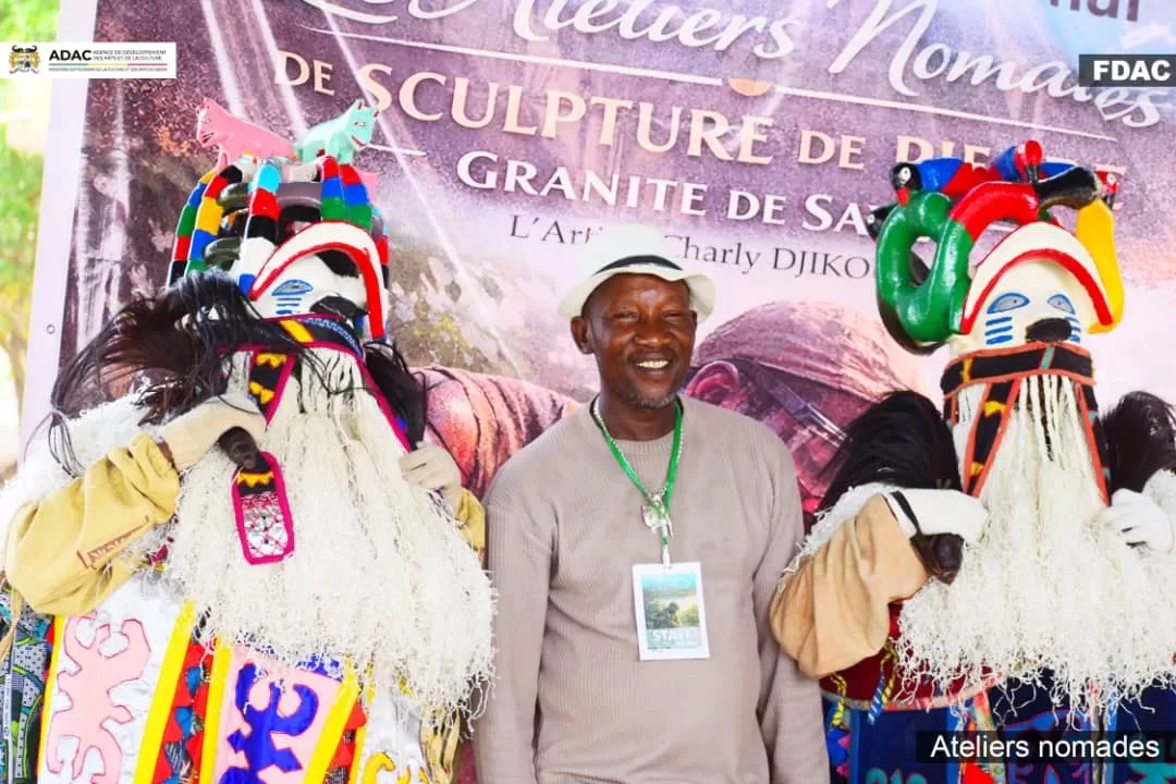 Un homme souriant pose entre deux personnes en costumes colorés et masques traditionnels devant une banderole événementielle