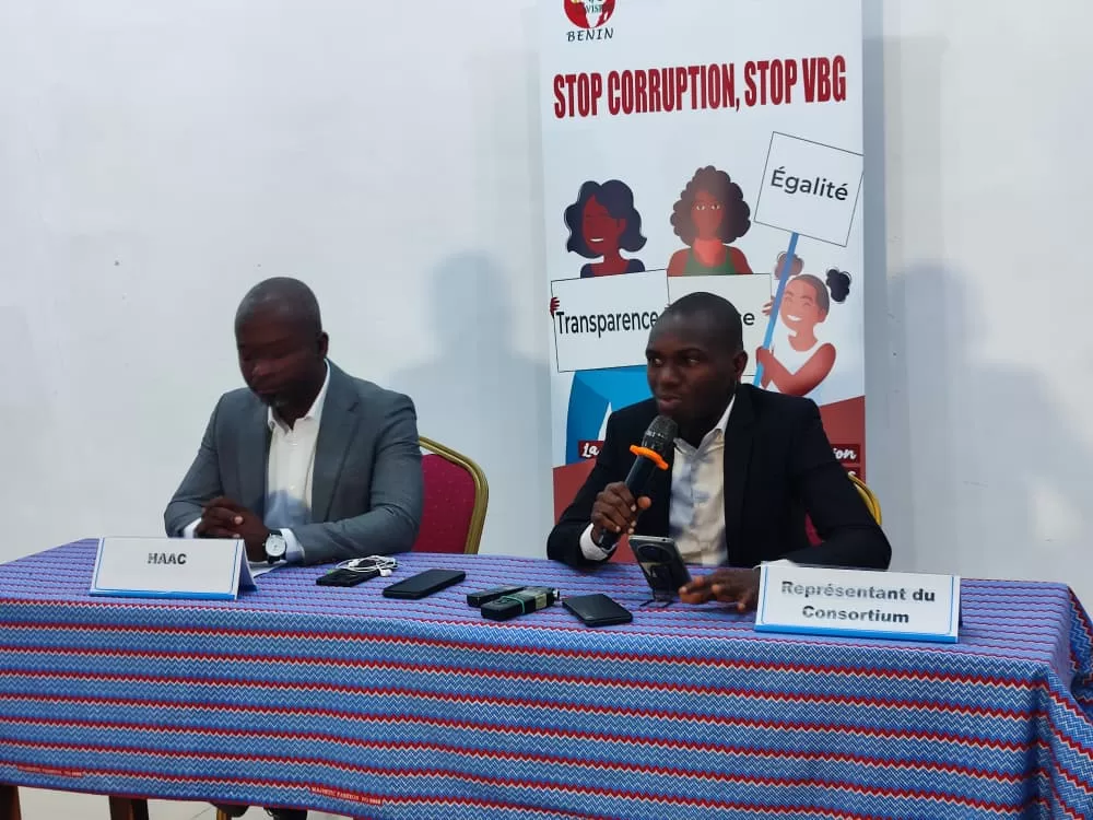 Deux hommes assis à une table avec micros et panneaux derrière eux sur le thème « Stop Corruption, Stop VBG