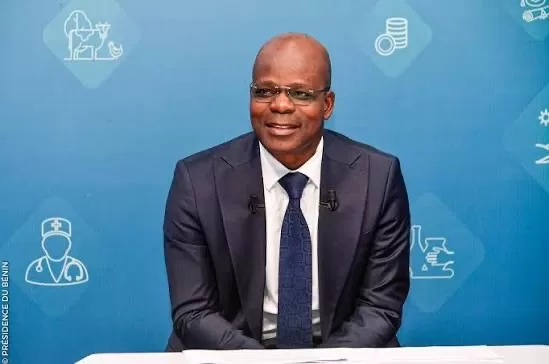 Homme en costume sombre et cravate assis devant un fond bleu avec des icônes blanches liées à la santé et l'économie